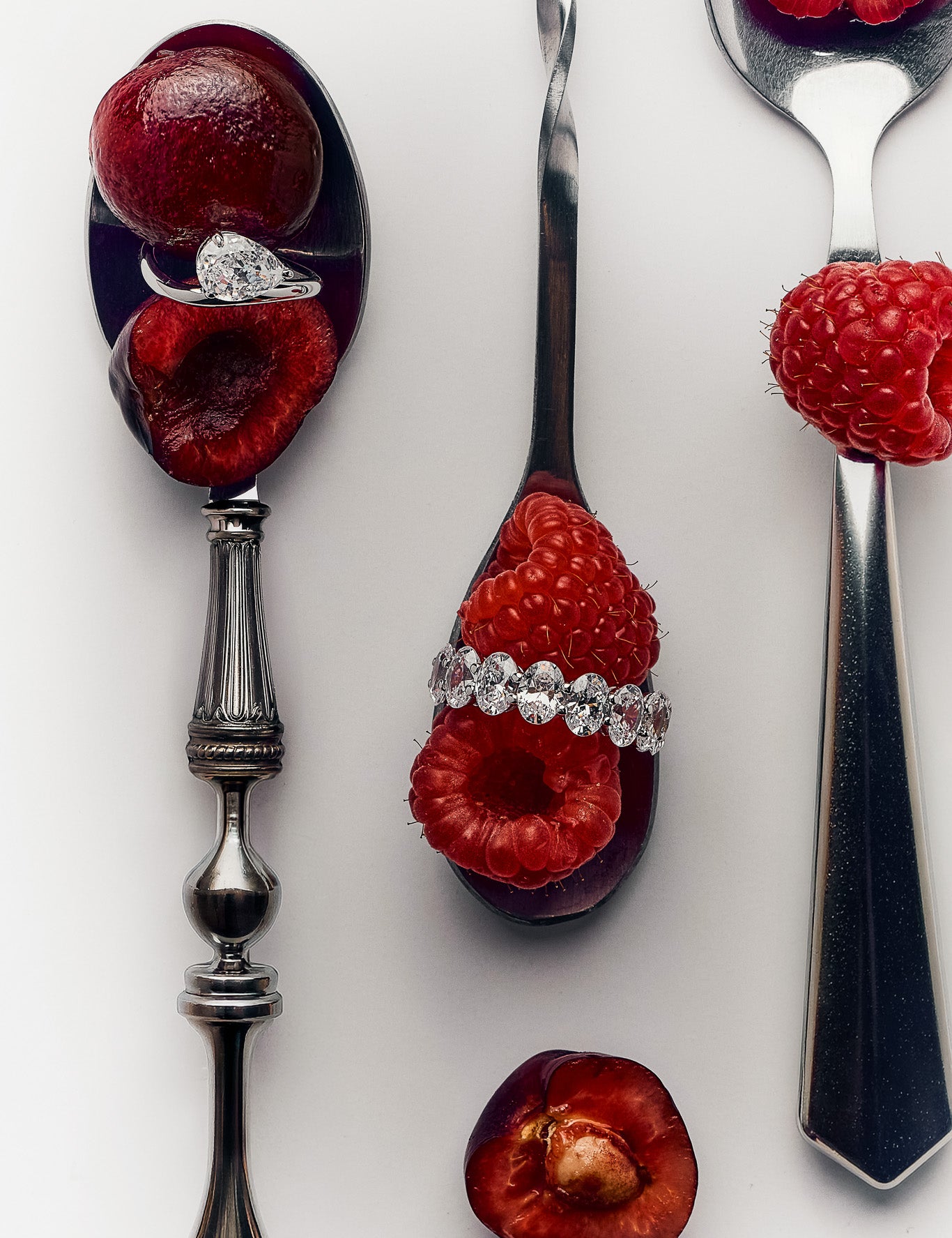 Three silver spoons arranged vertically on a white background, each holding a halved dark red plum or raspberry, with a lume hera ring - clear gemstone ring resting on the spoon bowl. A loose raspberry sits on the right spoon, and a halved plum is below.