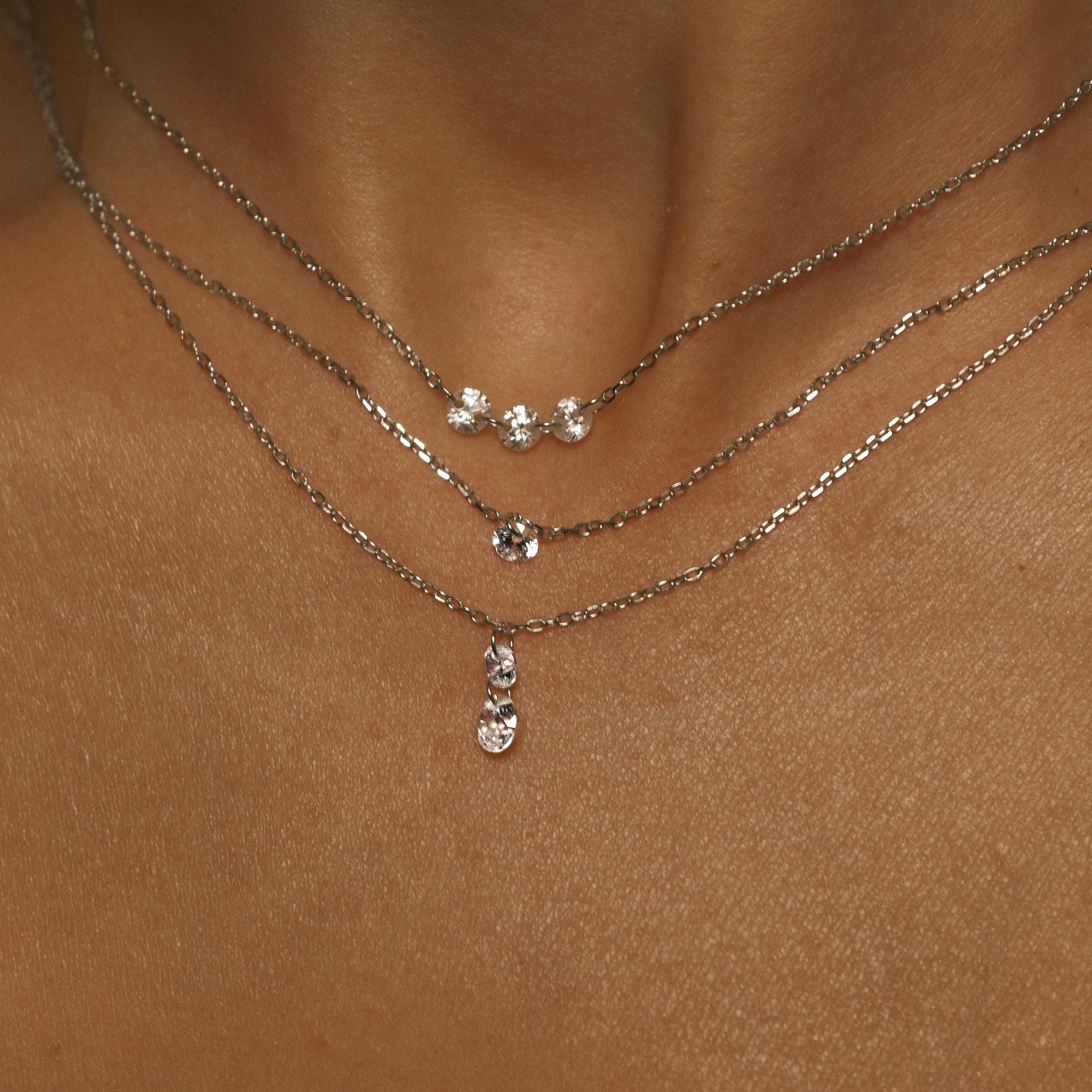 Close-up of a person wearing three silver necklaces with small pendants on a beige background.