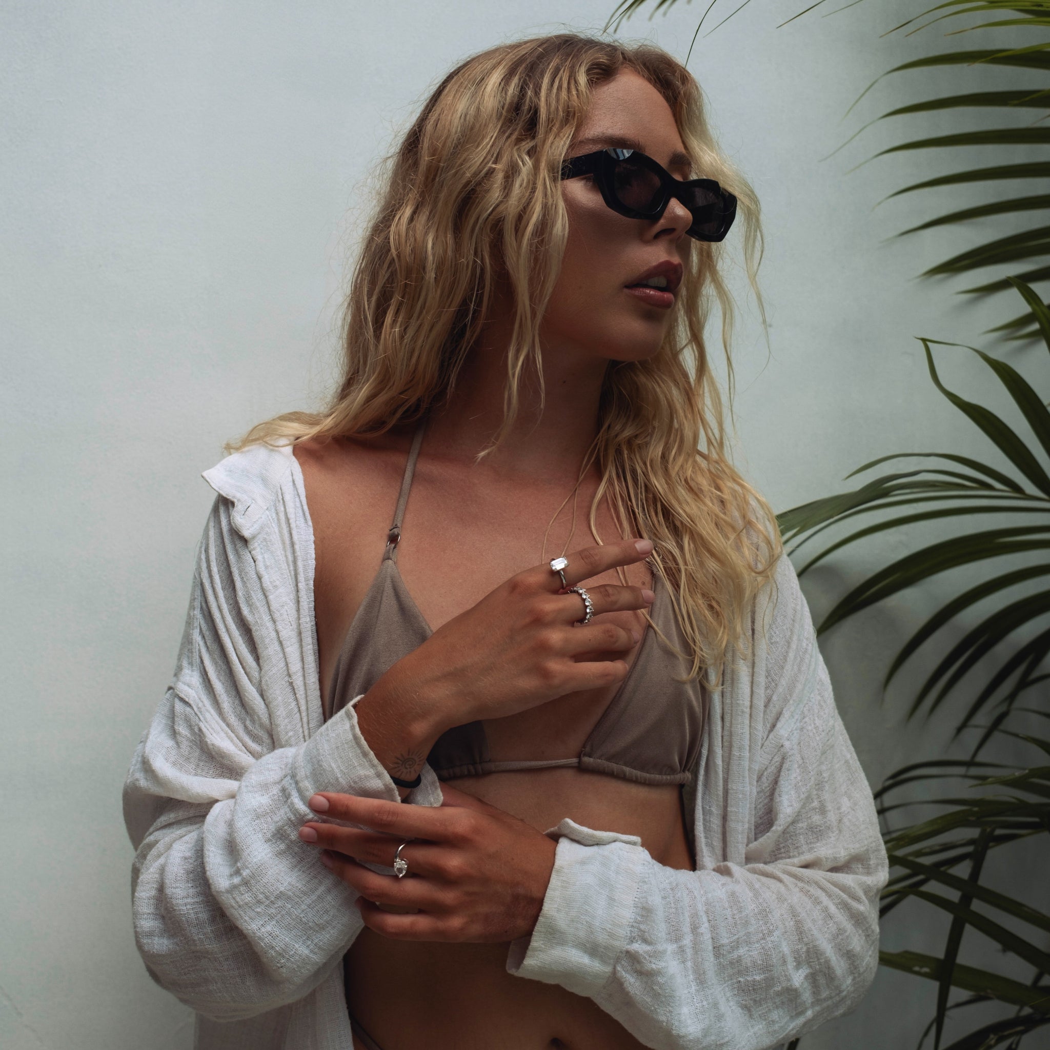 Woman wearing sunglasses and a white cardigan over a bikini, wearing lume jewellery's amore ring, roen ring and melora ring, standing against a light background with a plant.