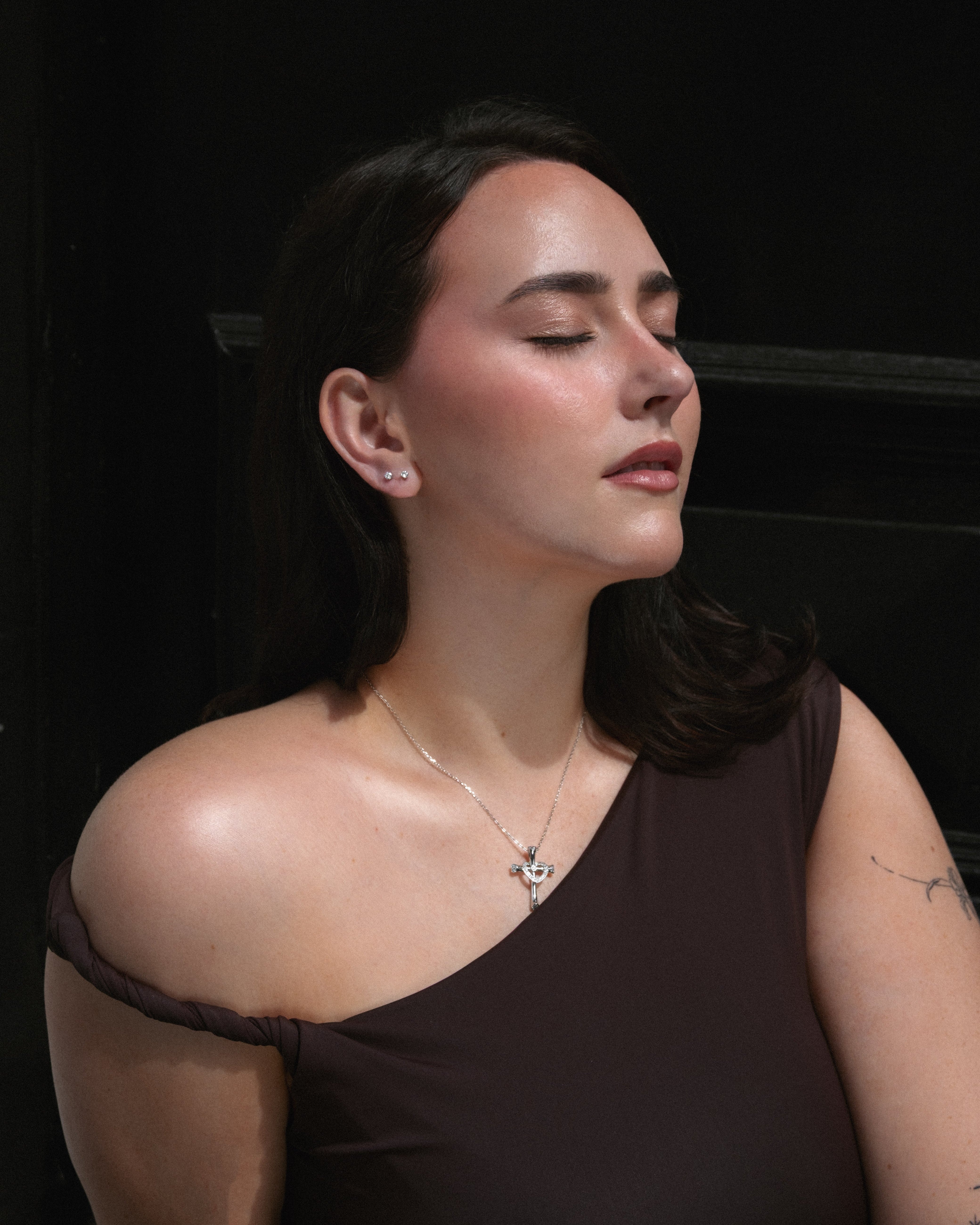 Woman wearing a brown one-shoulder top against a dark background, wearing the stella studs and silver necklace from lume jewellery