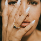 Close-up of a woman's face with one hand covering her eye, wearing the dues ring in gold and showing a tattoo.