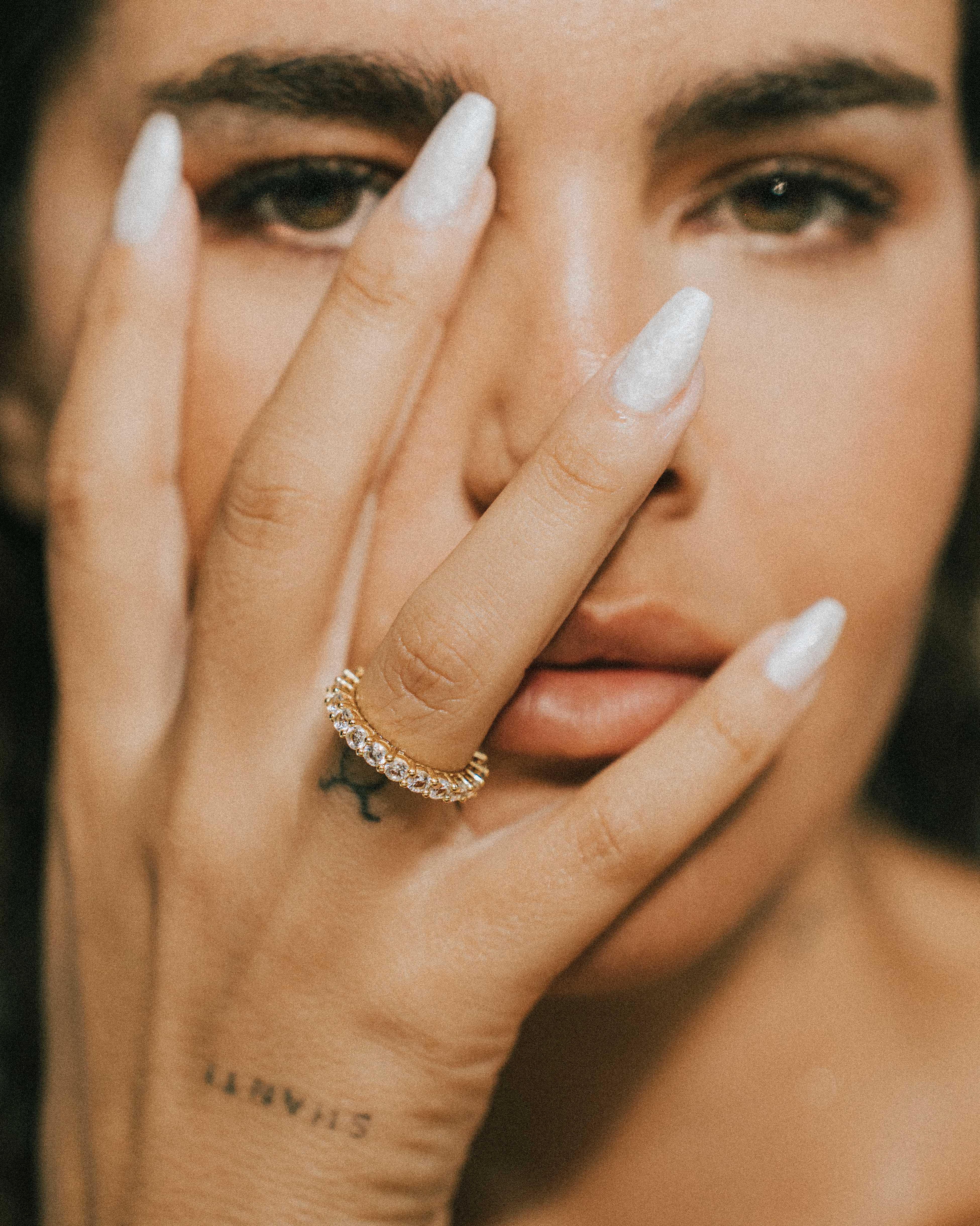 Close-up of a woman's face with one hand covering her eye, wearing the dues ring in gold and showing a tattoo.