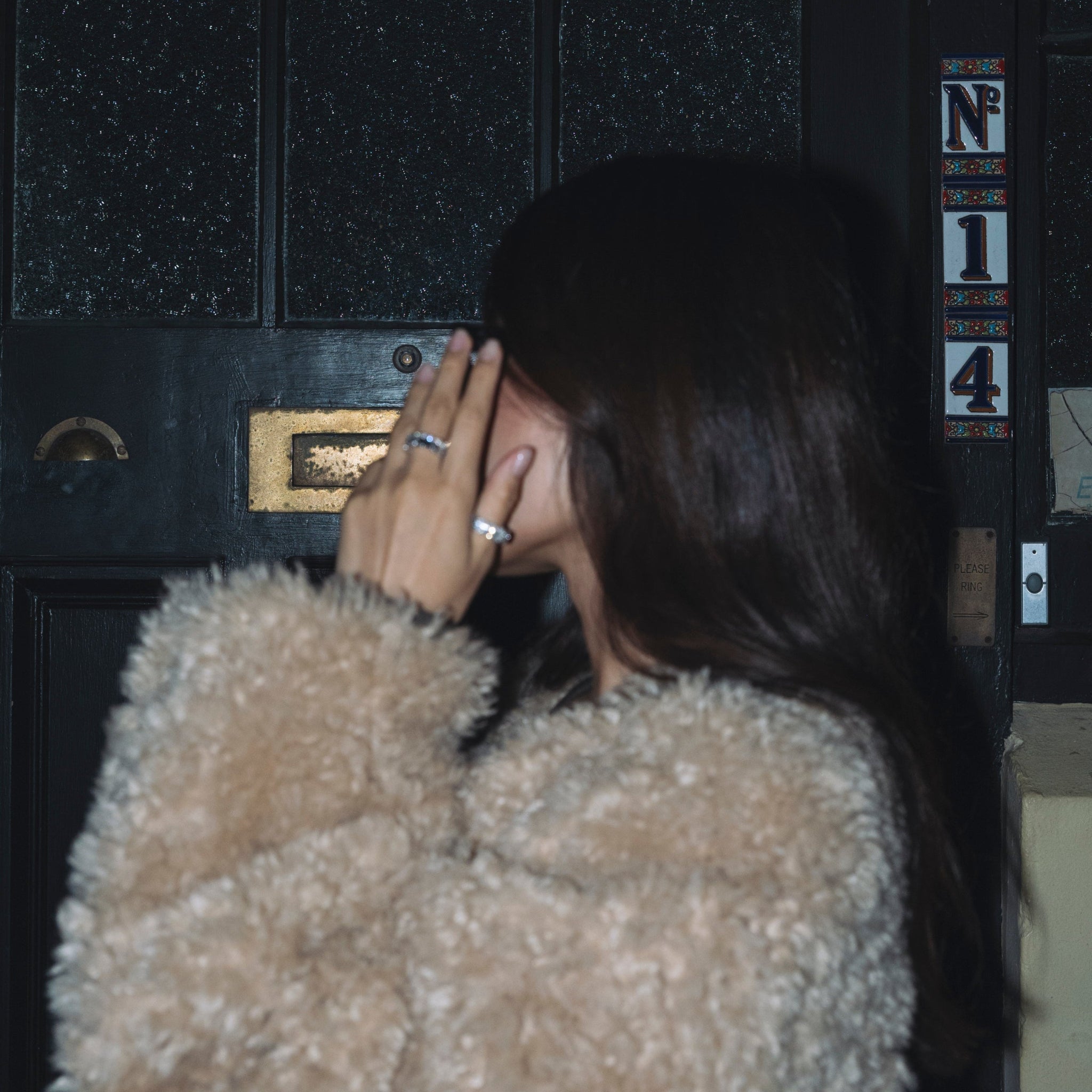 Person wearing a fluffy beige coat standing in front of a dark door, hand wearing lumejewellery's hera ring